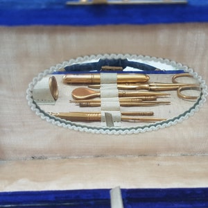 May include: A blue velvet jewelry box with a mirror inside. The mirror reflects a set of gold-toned manicure tools, including a nail file, tweezers, and a cuticle pusher.