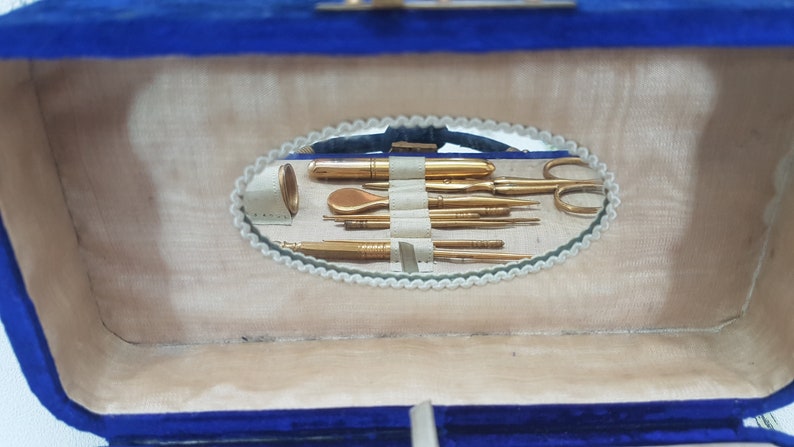 May include: A blue velvet jewelry box with a mirror inside. The mirror reflects a set of gold-toned manicure tools, including a nail file, tweezers, and a cuticle pusher.