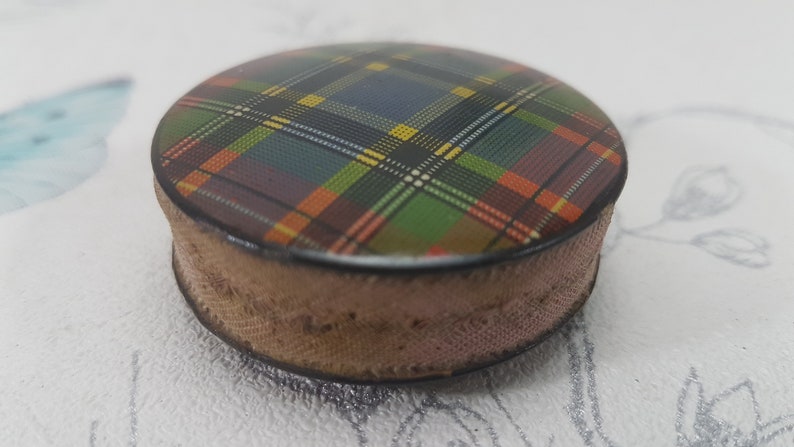 K&ouml;nnte beinhalten: Kleine, runde Schatulle mit einem Schottenmuster auf dem Deckel. Das Schottenmuster weist eine Mischung aus Farben auf, darunter Gr&uuml;n, Gelb, Orange und Blau. Die Seiten der Schatulle sind hellbraun.