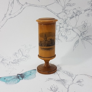 Puede incluir: Un recipiente cilíndrico de madera con tapa, que presenta una ilustración impresa en marrón oscuro de una iglesia y un paisaje urbano en el lateral. El objeto se apoya sobre una base circular y tiene un acabado liso en tono marrón claro.