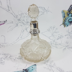 May include: A clear glass perfume bottle with a faceted design and a silver stopper. The bottle has a crystal-like stopper with a rounded top.