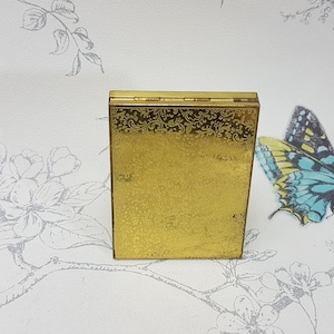May include: A gold compact with an ornate floral design. The compact is closed and has a hinged lid.