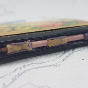 May include: A small black leather-bound notebook with a floral design on the cover. The notebook has a pink pencil attached to it with purple ribbon.
