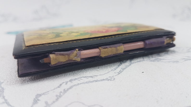 May include: A small black leather-bound notebook with a floral design on the cover. The notebook has a pink pencil attached to it with purple ribbon.