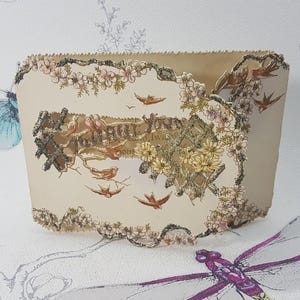 Puede incluir: Un recipiente de papel beige con un diseño floral y de pájaros. El recipiente está decorado con una ilustración detallada de un paisaje, pájaros y flores. Una mariposa azul y una libélula púrpura están en el fondo.