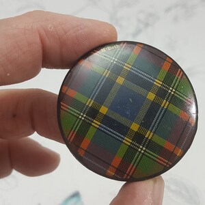 K&ouml;nnte beinhalten: Ein runder Knopf mit einem farbenfrohen Schottenmuster in Blau-, Gr&uuml;n-, Orange- und Gelbt&ouml;nen. Der Knopf hat eine gl&auml;nzende Oberfl&auml;che und einen dunkelbraunen Rand. Der Text "M. Beth" ist auf dem Knopf sichtbar.