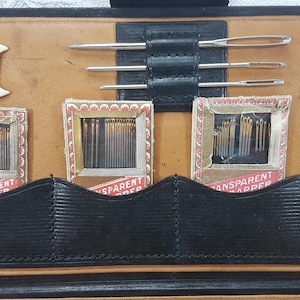 May include: A brown leather sewing kit with four compartments containing needles and thread. The kit has a black strap and a white spool holder.