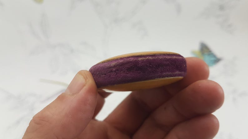 May include: A small, round, wooden object with a purple velvet center. The object is held in a hand.