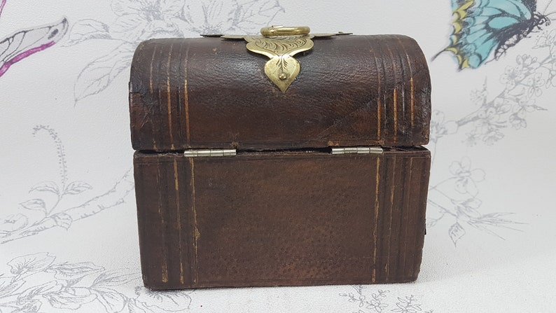 May include: A small, brown leather box with a brass latch and hinges. The box has a decorative design on the top and is closed.