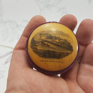 May include: A small, round, purple velvet box with a yellow lid. The lid has a black and white image of Edinburgh Castle with the text "Edinburgh Castle" printed below the image.