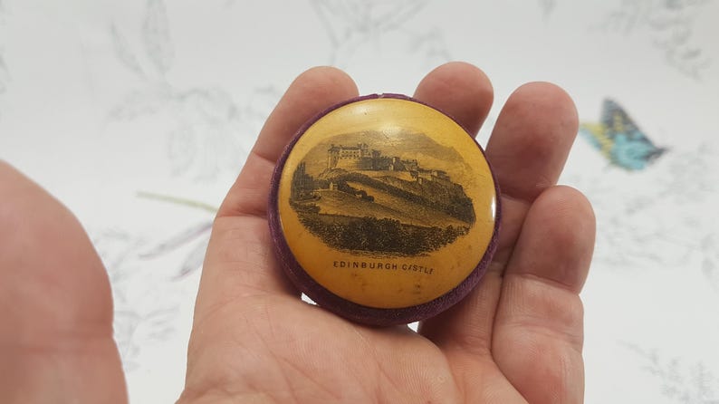 May include: A small, round, purple velvet box with a yellow lid. The lid has a black and white image of Edinburgh Castle with the text "Edinburgh Castle" printed below the image.