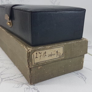 May include: A black leather box with a brass latch, sitting on top of a brown cardboard box with a handwritten label that reads "12 7/12 schr-17/12".