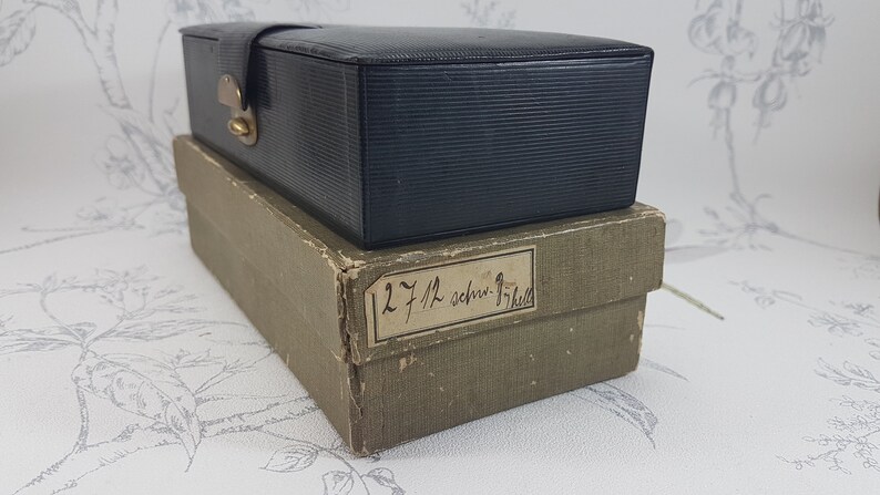 May include: A black leather box with a brass latch, sitting on top of a brown cardboard box with a handwritten label that reads "12 7/12 schr-17/12".