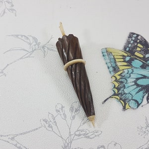 May include: A miniature wooden umbrella with a brown and white striped design. The umbrella is closed and has a white elastic band around it. The umbrella is sitting on a white surface with a floral pattern.