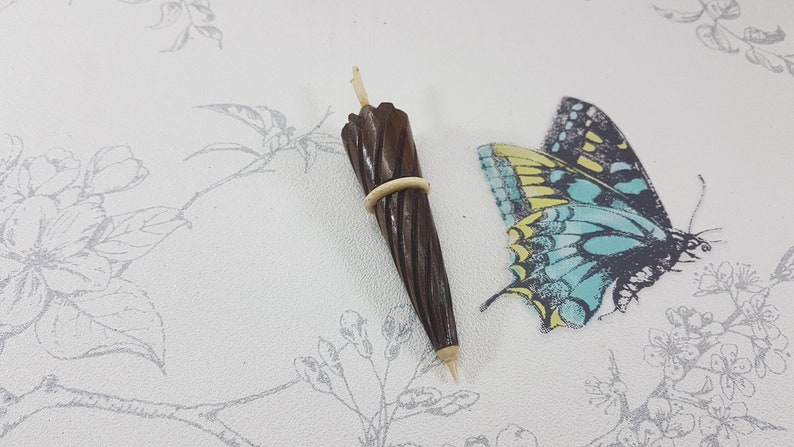 May include: A miniature wooden umbrella with a brown and white striped design. The umbrella is closed and has a white elastic band around it. The umbrella is sitting on a white surface with a floral pattern.