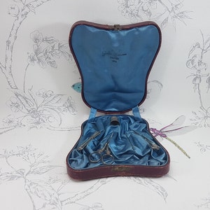May include: A vintage red leather sewing kit with blue satin lining. The kit contains a thimble, scissors, and two needles. The kit is open and the inside is visible. The kit has a label that reads "Guille Bros. Cutler, New York".