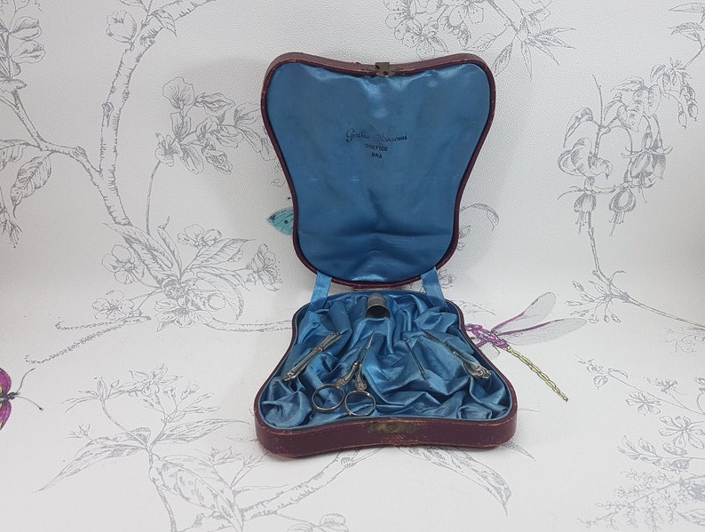 May include: A vintage red leather sewing kit with blue satin lining. The kit contains a thimble, scissors, and two needles. The kit is open and the inside is visible. The kit has a label that reads "Guille Bros. Cutler, New York".
