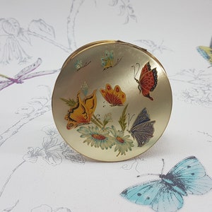 May include: A gold compact mirror with a floral design featuring butterflies in shades of yellow, orange, red, blue, and purple. The mirror is surrounded by a white background with a floral pattern.