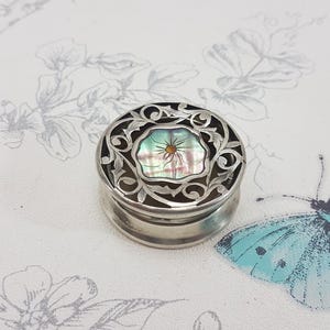 Pode incluir: Uma caixa de joias de prata com um design floral intrincado e uma incrustação de madrepérola. A caixa está fechada e está sobre uma superfície branca com um padrão floral.