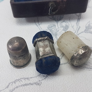 May include: Three silver thimbles, one with a blue velvet top, one with a white top, and one with a silver top.