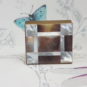 May include: A square box with a geometric pattern made of mother of pearl and brass. The box is light brown in colour and decorated with a white and brown pattern.