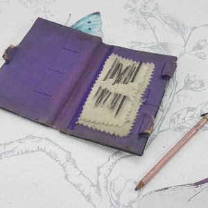 May include: A small purple leather needle book with a white fabric interior and a set of needles inside. The book is open and the needles are visible. A pink pencil is lying on a white surface with a floral pattern.