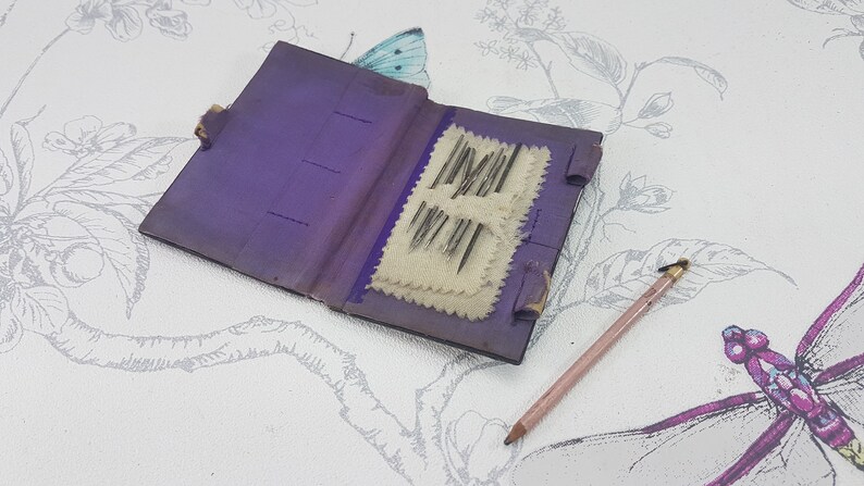 May include: A small purple leather needle book with a white fabric interior and a set of needles inside. The book is open and the needles are visible. A pink pencil is lying on a white surface with a floral pattern.