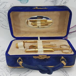May include: A blue velvet travel case with gold hardware and a mirror inside. The case contains a set of gold-toned manicure tools, including a nail file, cuticle pusher, tweezers, and scissors.
