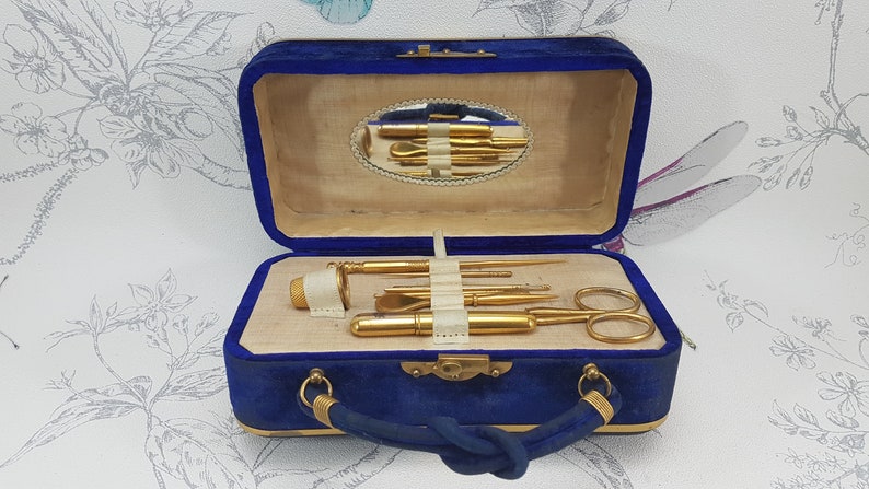 May include: A blue velvet travel case with gold hardware and a mirror inside. The case contains a set of gold-toned manicure tools, including a nail file, cuticle pusher, tweezers, and scissors.