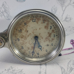 May include: A silver clock with a white face and black hands. The clock has a round face and a silver case. The clock is set to 10:50.