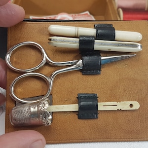 May include: A brown leather sewing kit with a silver thimble, scissors, and two bone needle threaders. The kit is open and the tools are visible.