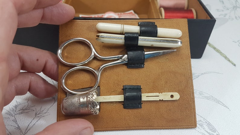 May include: A brown leather sewing kit with a silver thimble, scissors, and two bone needle threaders. The kit is open and the tools are visible.
