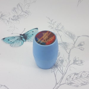 Op de afbeelding: Een kleine blauwe tonvormige container met een kleurrijk patroon aan de bovenkant. Het patroon heeft een rood, geel en blauw ruitpatroon met een gouden distel.