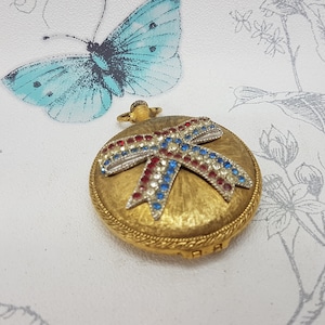 Op de afbeelding: Een goudkleurig compact met een rode, witte en blauwe juwelen strik. Het compact heeft een getextureerd oppervlak en een decoratieve rand.