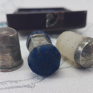 May include: Three silver thimbles, one with a blue velvet cushion, one with a clear glass top, and one with a silver top. The thimbles are all decorated with intricate floral patterns.