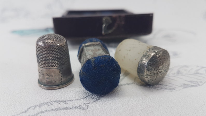 May include: Three silver thimbles, one with a blue velvet cushion, one with a clear glass top, and one with a silver top. The thimbles are all decorated with intricate floral patterns.