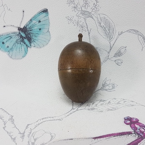 Puede incluir: Un recipiente de madera en forma de bellota, de color marrón, con un pequeño tallo en la parte superior. Se encuentra sobre una superficie blanca decorada con una mariposa azul claro y motivos florales.