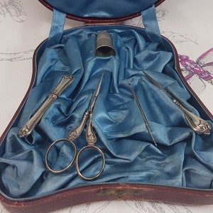 May include: A vintage sewing kit in a red velvet case with a blue satin lining. The kit includes a pair of silver scissors, two silver needle holders, a thimble, and a needle.
