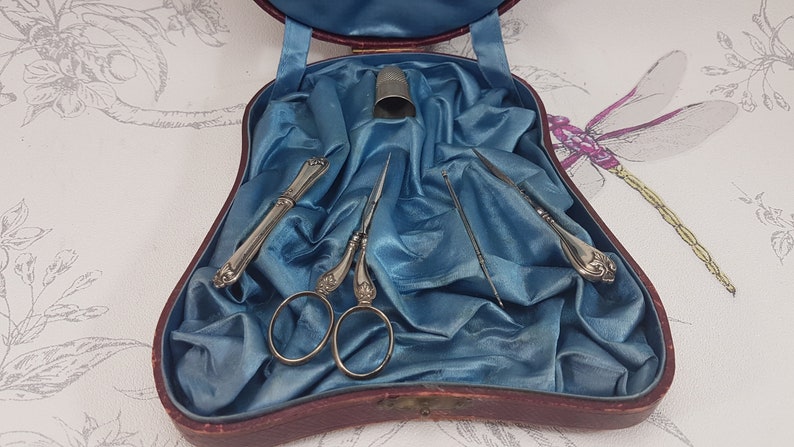 May include: A vintage sewing kit in a red velvet case with a blue satin lining. The kit includes a pair of silver scissors, two silver needle holders, a thimble, and a needle.
