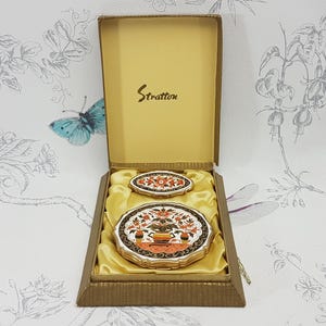 以下が含まれることがあります： 金色の箱に入ったストラトン製のコンパクトセット。コンパクトは白い背景にオレンジ、赤、緑の色の花柄が描かれている。箱には金色の文字で「ストラトン」と印刷されている。