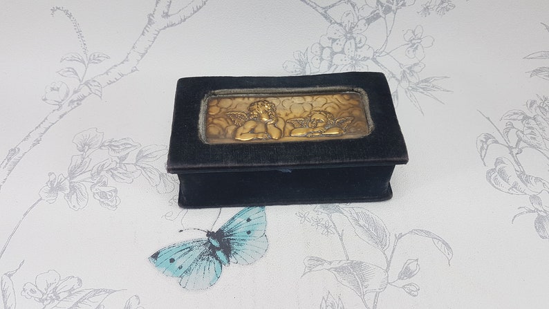 May include: A small black velvet box with a gold metal cherub design on the lid.