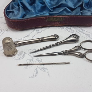 May include: A vintage sewing kit containing silver-toned tools, including a thimble, two needle threaders, a pair of scissors, and a needle. The tools are arranged on a white surface with a floral pattern.
