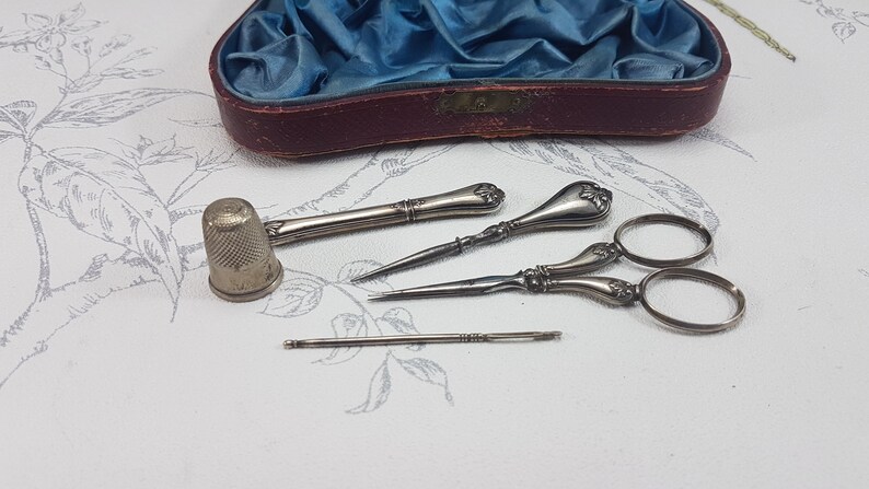 May include: A vintage sewing kit containing silver-toned tools, including a thimble, two needle threaders, a pair of scissors, and a needle. The tools are arranged on a white surface with a floral pattern.