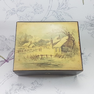 Peut inclure: Une boîte en bois avec un couvercle à charnières. Le couvercle est décoré d'une illustration vintage d'une scène d'hiver enneigée avec deux maisons et une clôture. Les maisons ont des toits enneigés et des fenêtres.