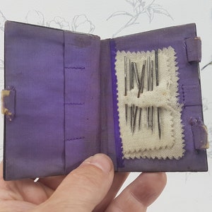 May include: A small purple velvet book with a flap closure, open to reveal a white fabric pocket containing a collection of sewing needles.