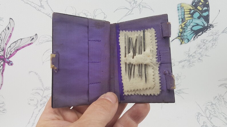 May include: A small purple velvet book with a flap closure, open to reveal a white fabric pocket containing a collection of sewing needles.