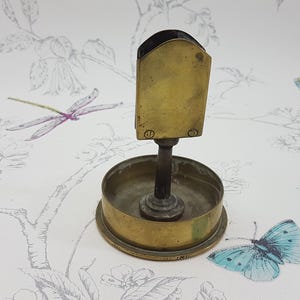 Puede incluir: Un encendedor de cigarrillos de latón con una base redonda y una parte superior rectangular. El encendedor tiene una pequeña perilla redonda en el lateral.