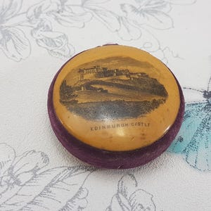 May include: A small, round, purple velvet box with a cream-coloured lid. The lid has a black and white image of Edinburgh Castle with the text "EDINBURGH CASTLE" printed on it.