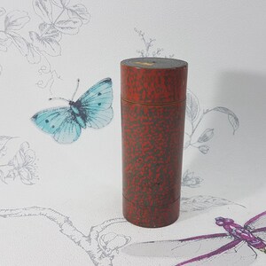Op de afbeelding: Een cilindrische container met een rode gespikkelde afwerking. De container staat rechtop tegen een achtergrond van een vlinder en bloemmotieven. De container wordt waarschijnlijk gebruikt voor opslag.