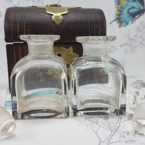 May include: Two clear glass bottles with stoppers in a wooden box with brass accents. The bottles are square shaped and have a clear liquid inside. The box is closed and has a brass latch.